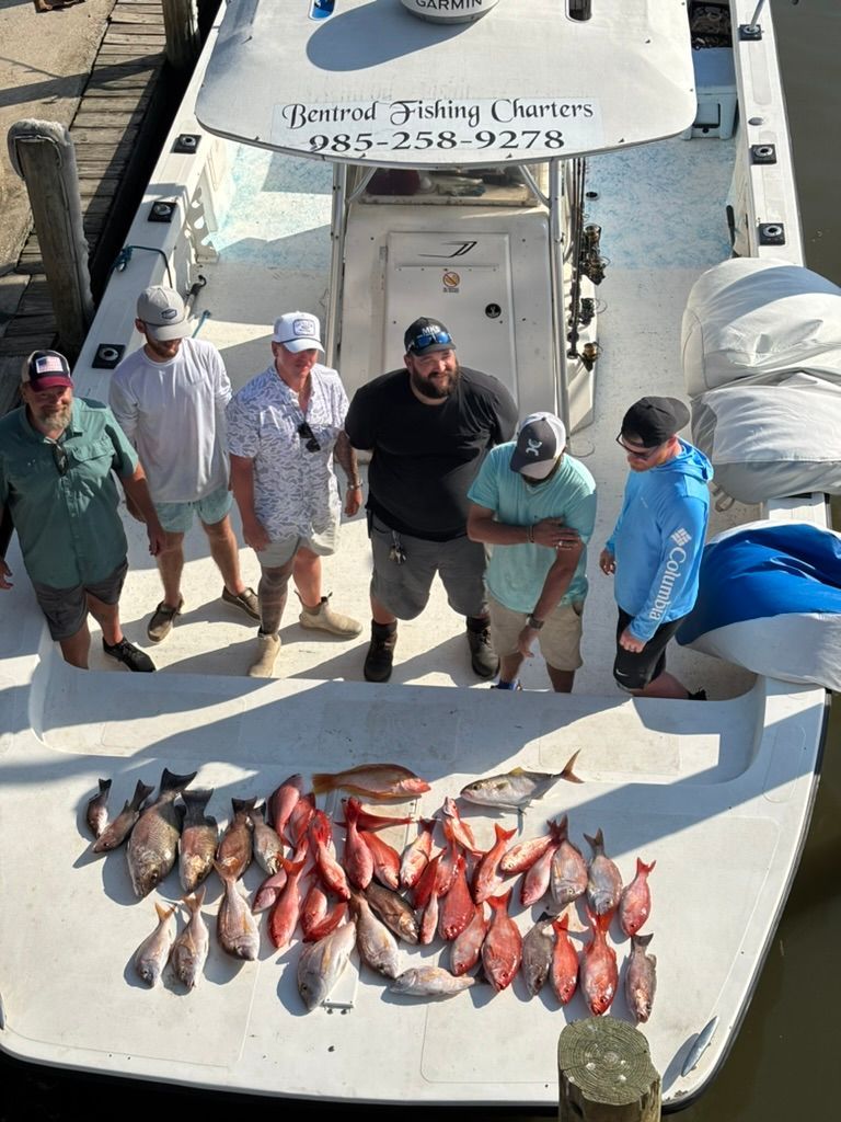 Six people stand on a boat deck behind a large catch of colorful fish, with a charter business sign on the console.