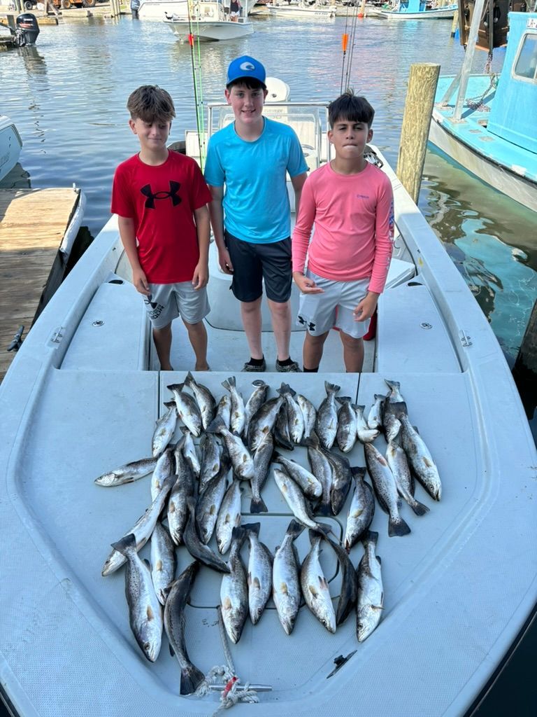 Three people stand on a boat deck behind a large catch of fish in a harbor.