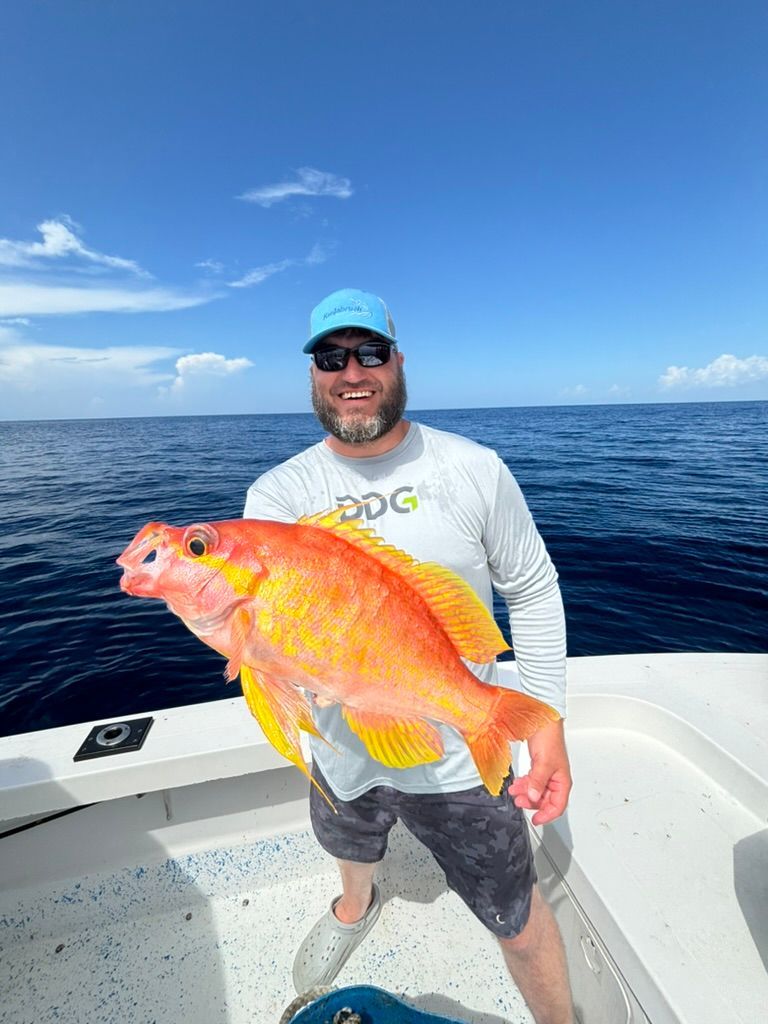 A smiling angler stands on a boat on the ocean, holding a large, bright orange fish.