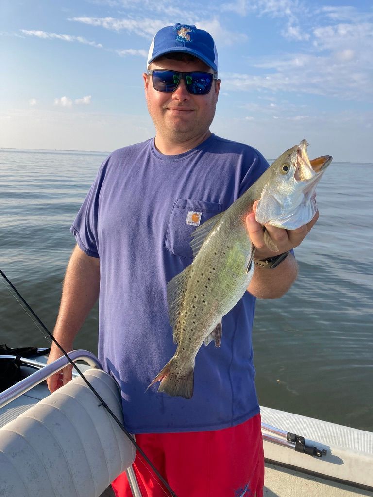 A person in a blue t-shirt and baseball cap stands on a boat, holding up a large spotted seatrout over calm water.