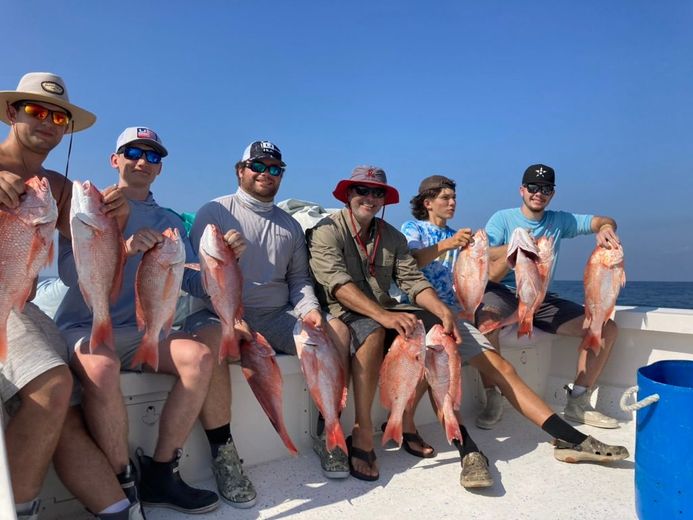 Six people sit on a boat deck wearing sunglasses and hats, each holding a red snapper fish against a bright blue sky.