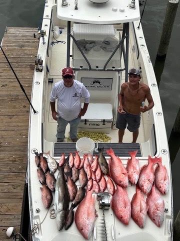 Two people stand on a boat at a dock behind a large catch of fresh red snapper and a grouper arranged on the deck.