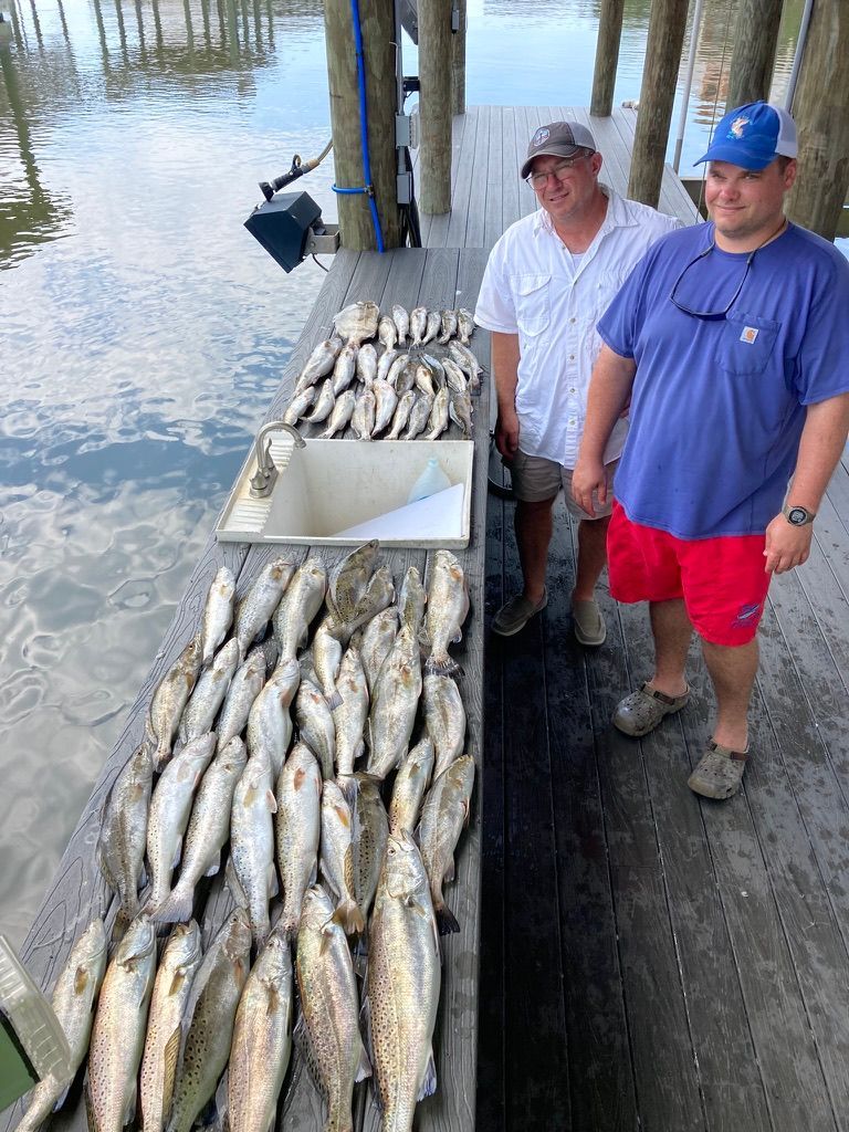Two men stand on a dock by water, beside a large catch of fish arranged on the wooden surface.