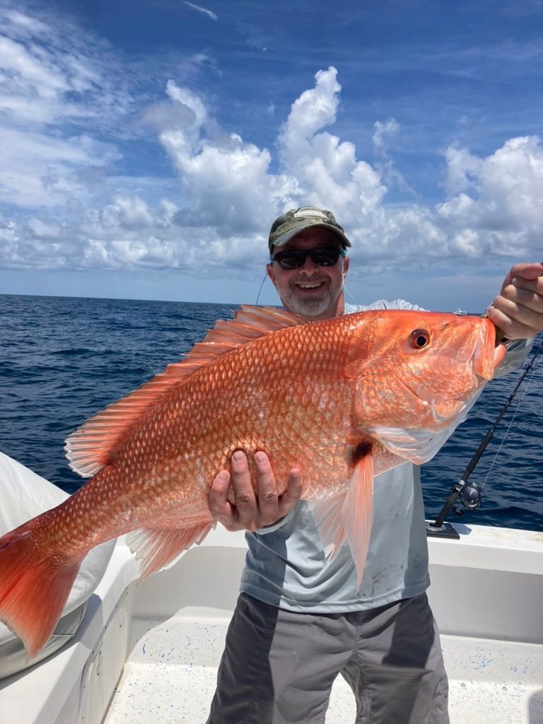 A person wearing sunglasses and a hat holds a large, vibrant red snapper fish on a boat against a blue sky and ocean.