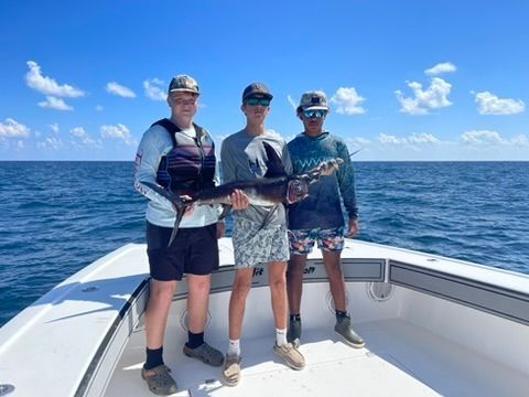 Three people holding a swordfish on the bow of a boat out on the blue ocean under a clear sky.