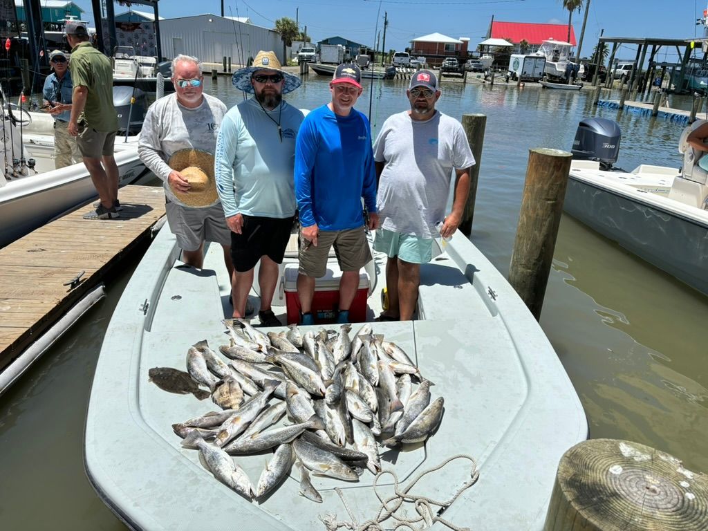 Four people stand on a boat at a marina with a large catch of fish spread out on the deck in front of them.