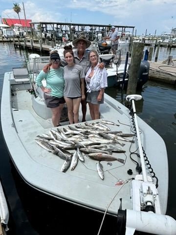 Four people stand on a boat at a marina with a large catch of fish laid out on the deck in front of them.