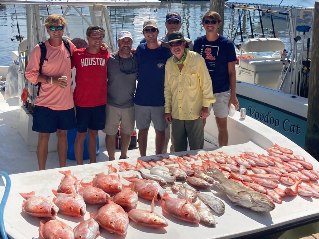 Seven people stand on a boat deck behind a large table filled with their catch of many red snapper and one larger fish.