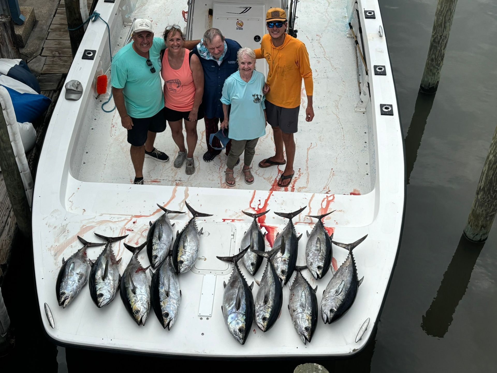Five people stand on a boat deck behind a large catch of fresh tuna fish at a dock.