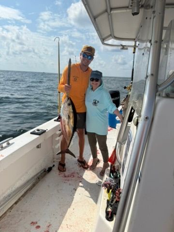 Two people stand on a boat deck holding a freshly caught tuna against a sunny ocean background.