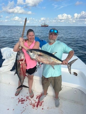 Two people stand on a boat deck holding freshly caught tuna fish against a blue sea background.
