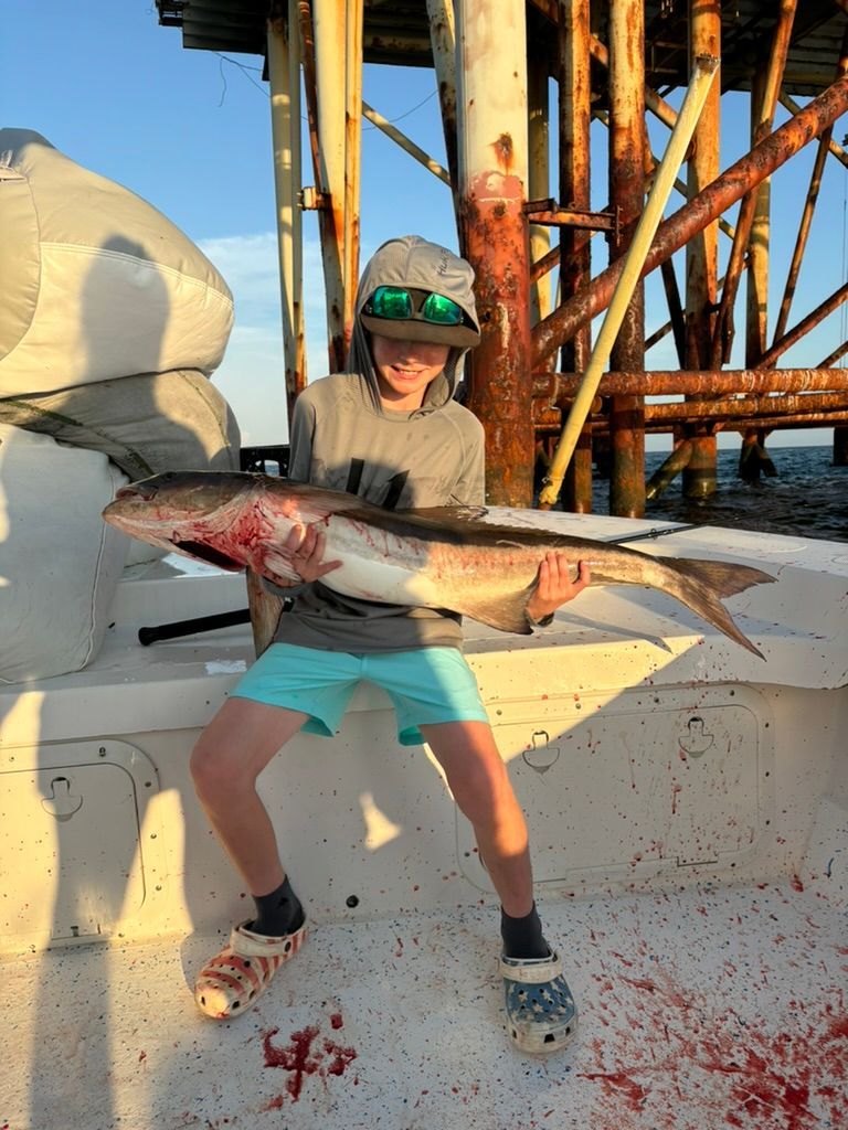 A person wearing a hat and sunglasses holds a large fish on a boat deck in front of an offshore oil platform.