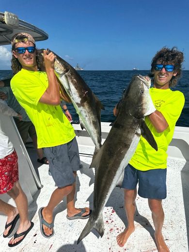 Two people in neon yellow shirts holding large fish on a boat deck under a clear blue sky.