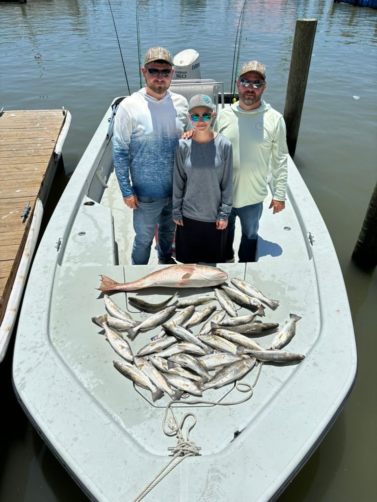 Three people stand in a boat moored at a dock with a large catch of fresh fish spread out on the deck.