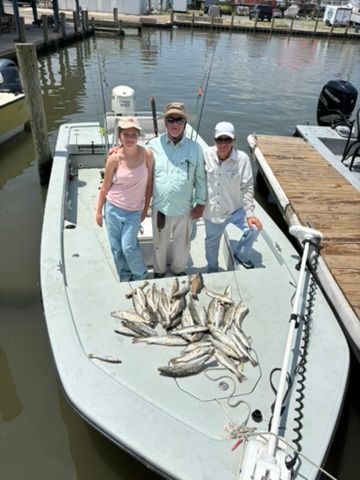Three people stand on a boat docked at a pier with their catch of many fish laid out on the deck.