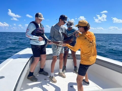 Four people on a boat deck hold a caught fish over the water on a sunny day.