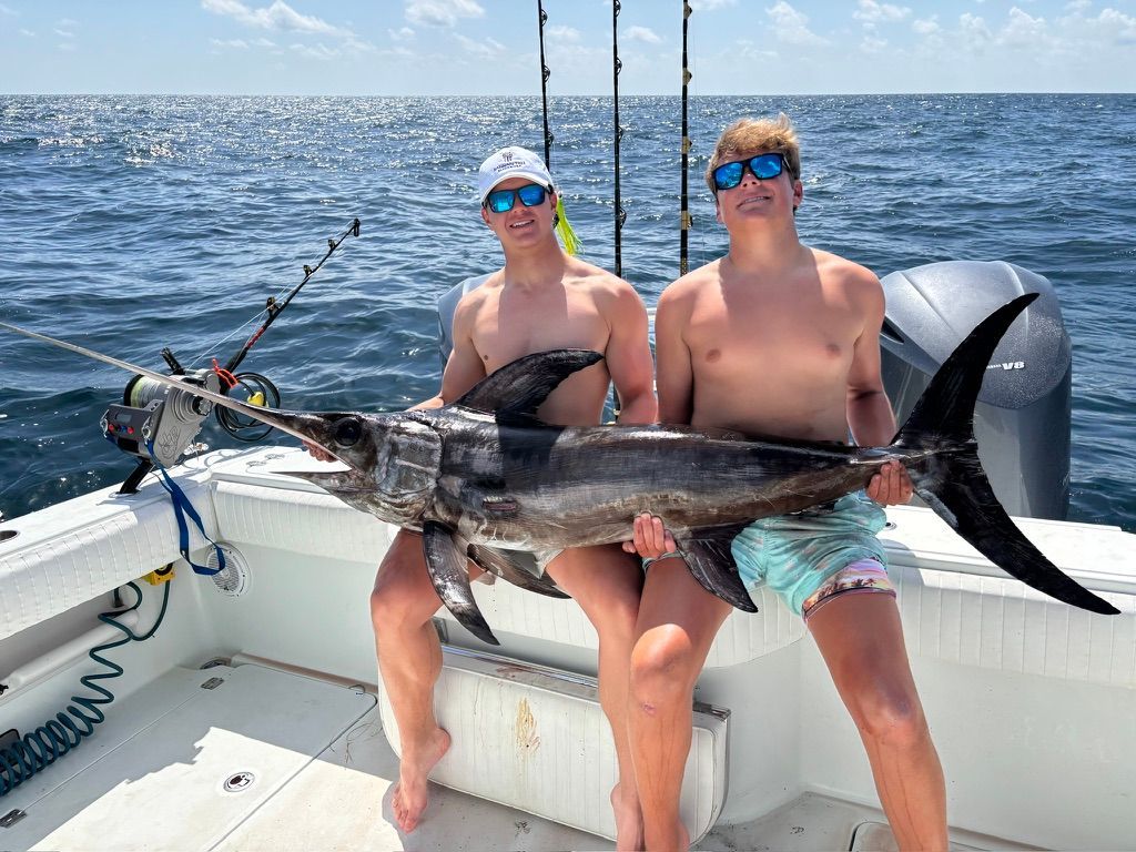 Two shirtless individuals sit on a boat deck, smiling while holding a large, caught fish between them.