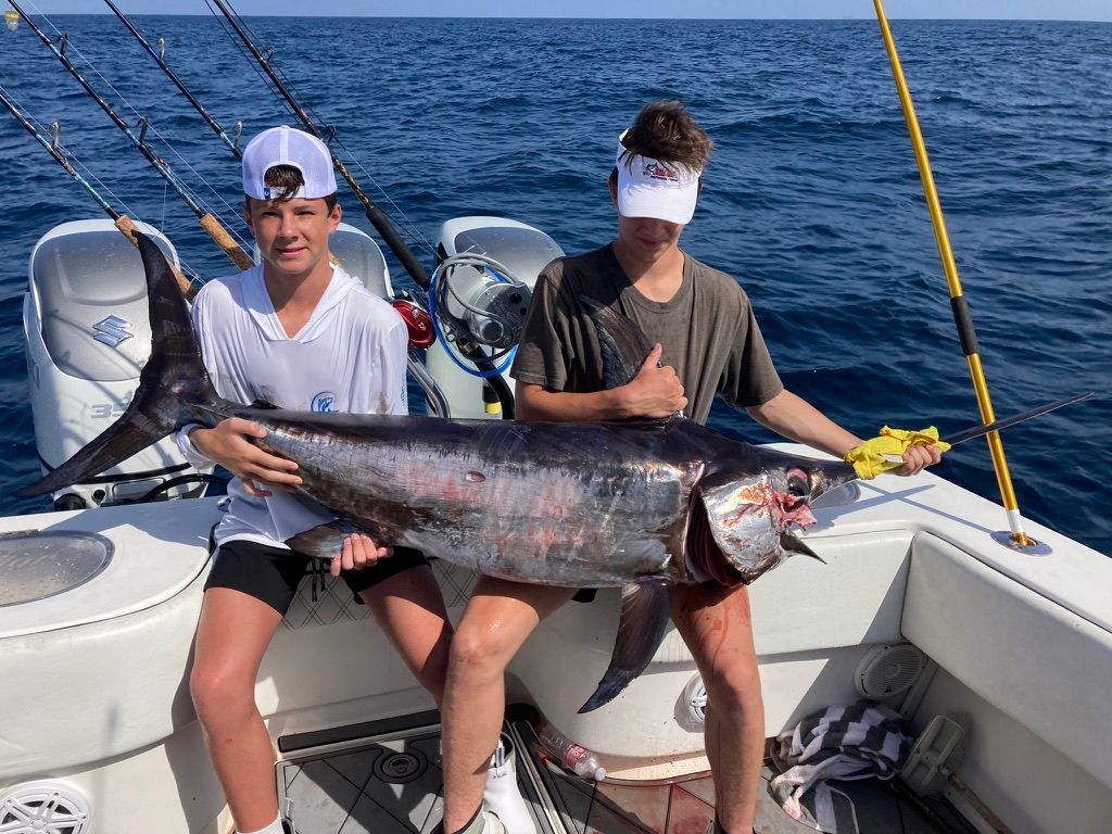Two people on a boat deck hold a large, dark-colored billfish horizontally across their laps against a blue ocean.