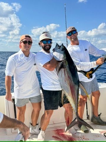 Three people wearing caps and sunglasses pose on a boat deck, holding a large caught tuna between them.