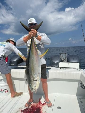 A person on a boat holds a large yellowfin tuna by its tail, with another person visible on the deck nearby.