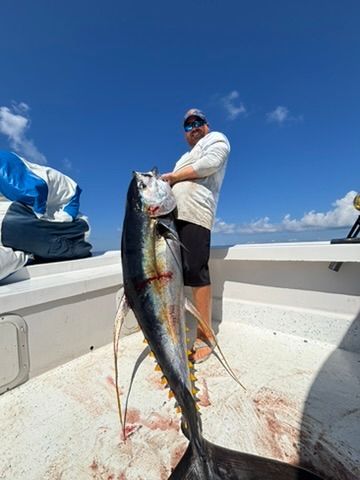 A person stands on a boat deck holding a large, freshly caught tuna against a clear blue sky.