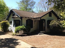 A small white house with a brown roof is surrounded by trees and bushes.