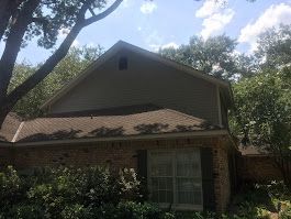 A house with a roof that is covered in shingles is surrounded by trees and bushes.