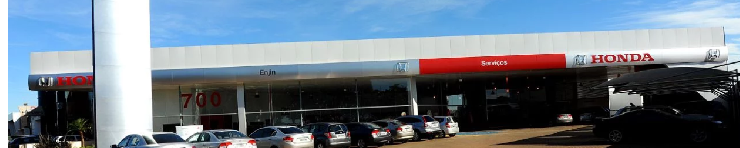 Lojas de um centro comercial com toldos vermelhos e carros estacionados sob um céu azul.