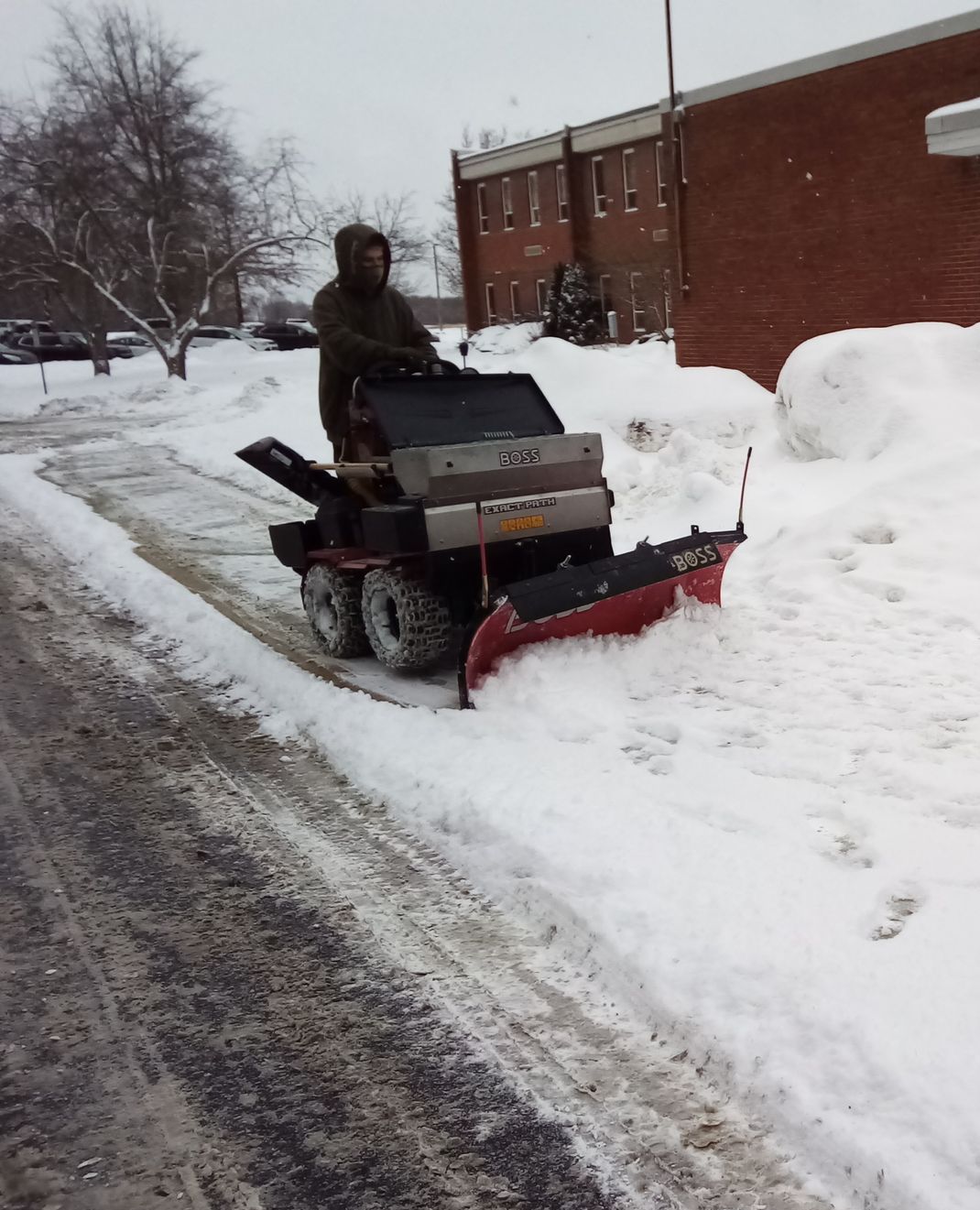 Person on a red snowblower clearing snow beside a brick building and road