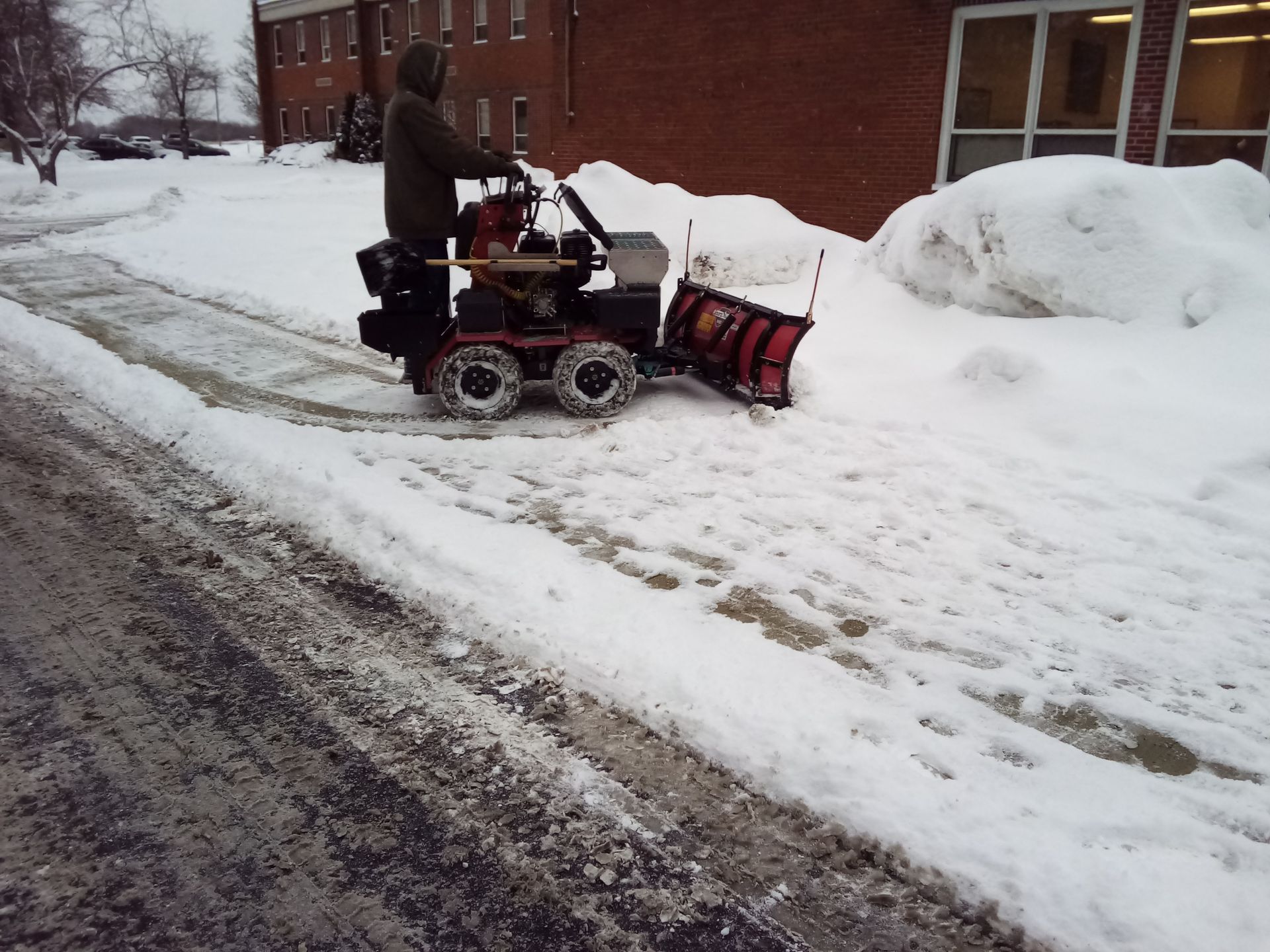 A man is pushing a snow plow down a snowy sidewalk