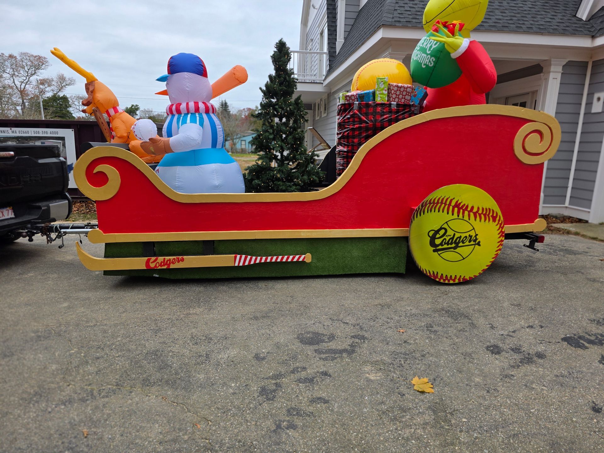 A red Santa sleigh float, decorated with baseball bats and balls, features a snowman and Grinch figure on a driveway.