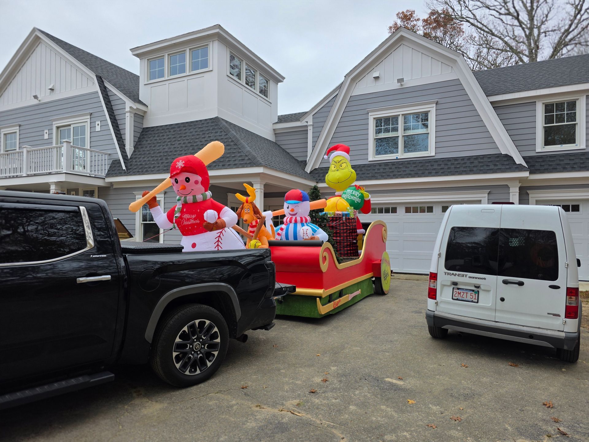 A black pickup truck tows a trailer with Grinch and snowman inflatable holiday decorations in a residential driveway.