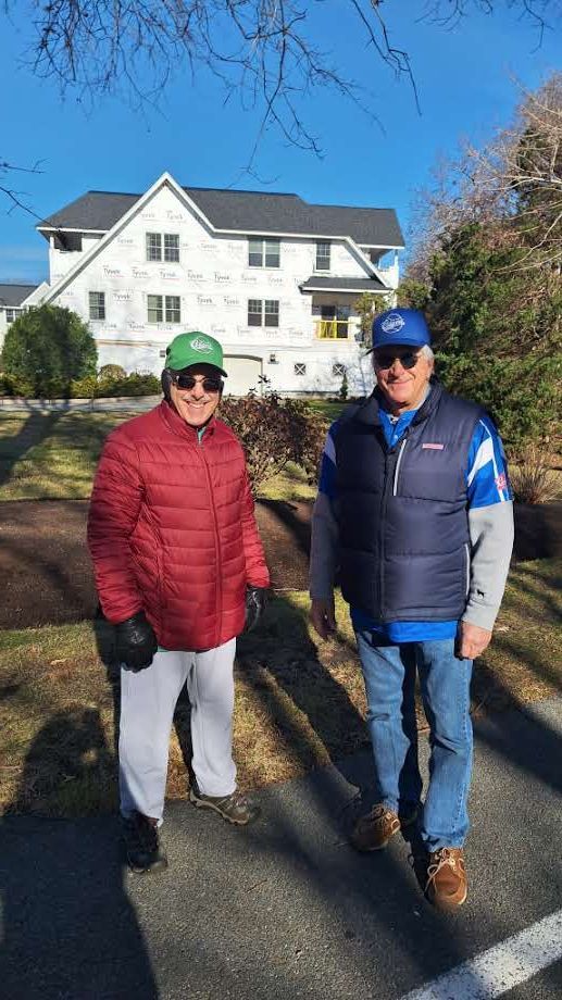 Two men in caps, jackets, and sunglasses stand outside on a sunny day in front of a white house under construction.