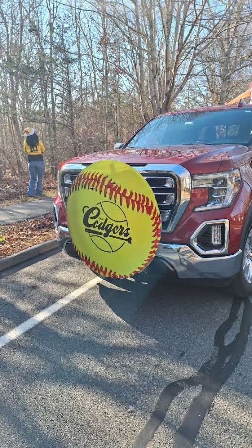 A maroon pickup truck with a large, yellow softball-themed sign attached to its front grille, parked in an outdoor lot.