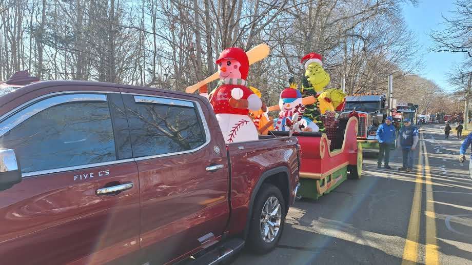 A maroon pickup truck pulls a decorated sleigh with a baseball-themed snowman and The Grinch on a sunny road.