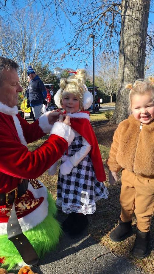 Santa Claus crouches to talk to a child in a red cape and buffalo plaid dress, while another child stands nearby.