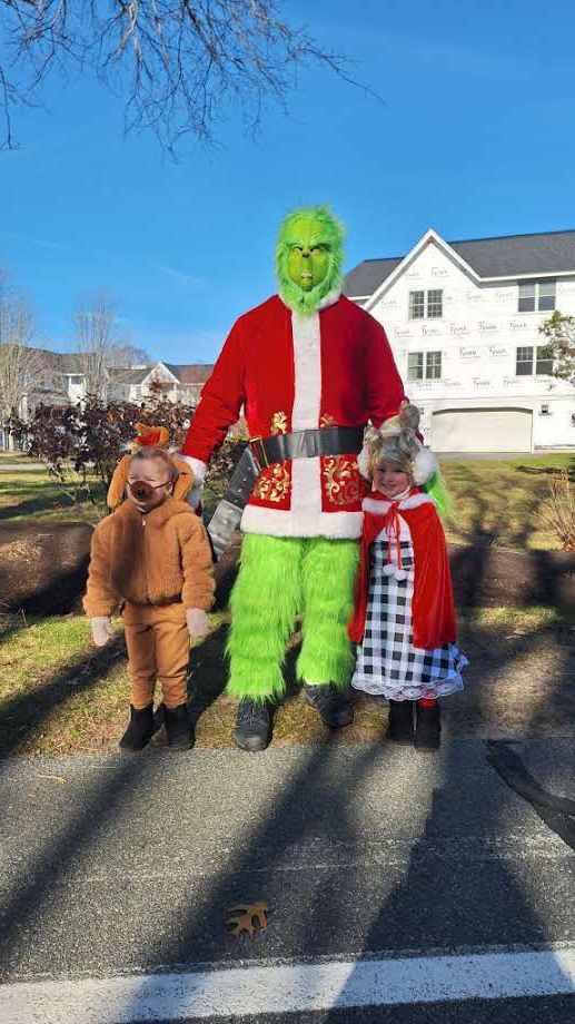 A person in a Grinch costume, dressed as Santa, stands outdoors between a child in a reindeer suit and one in a dress.