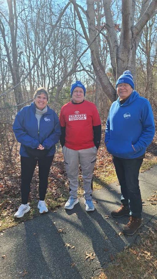 Three people stand on a paved path in a wooded area, wearing casual athletic clothing and knit hats.