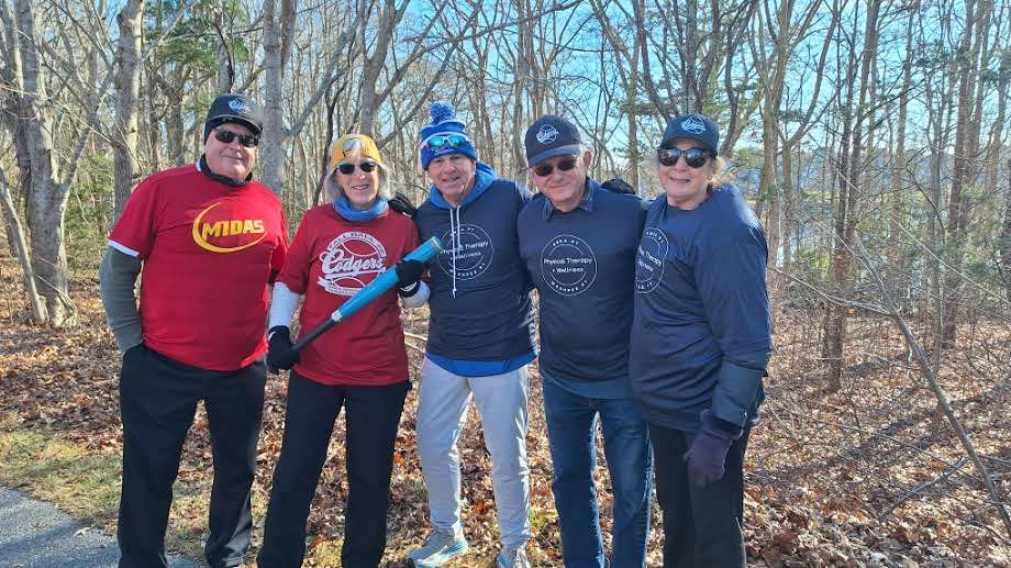 Five people in casual clothing and caps stand together outdoors in a wooded area, one holding a blue baseball bat.