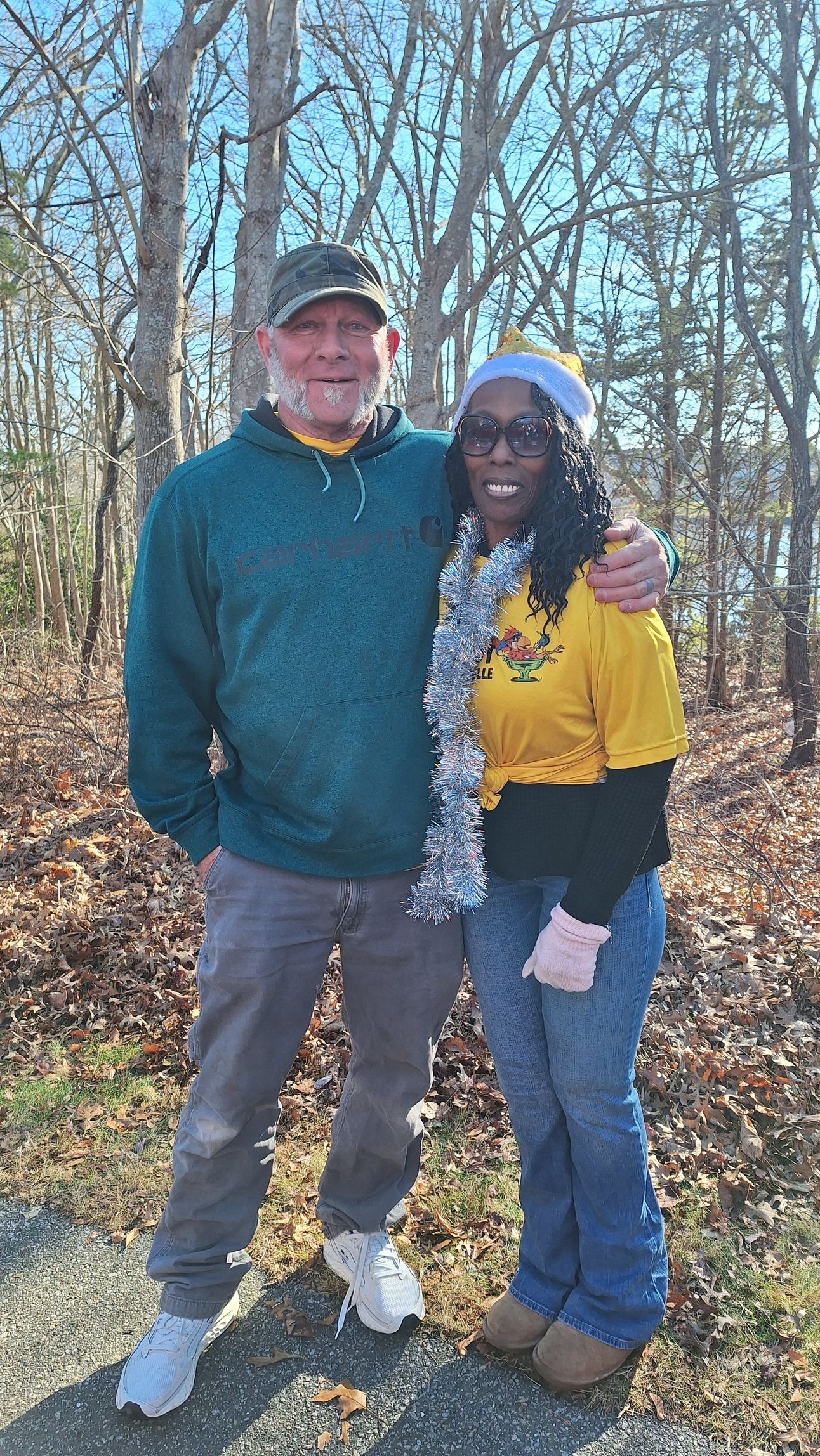 A couple stands smiling in a wooded area with autumn leaves on the ground, wearing casual outdoor attire.