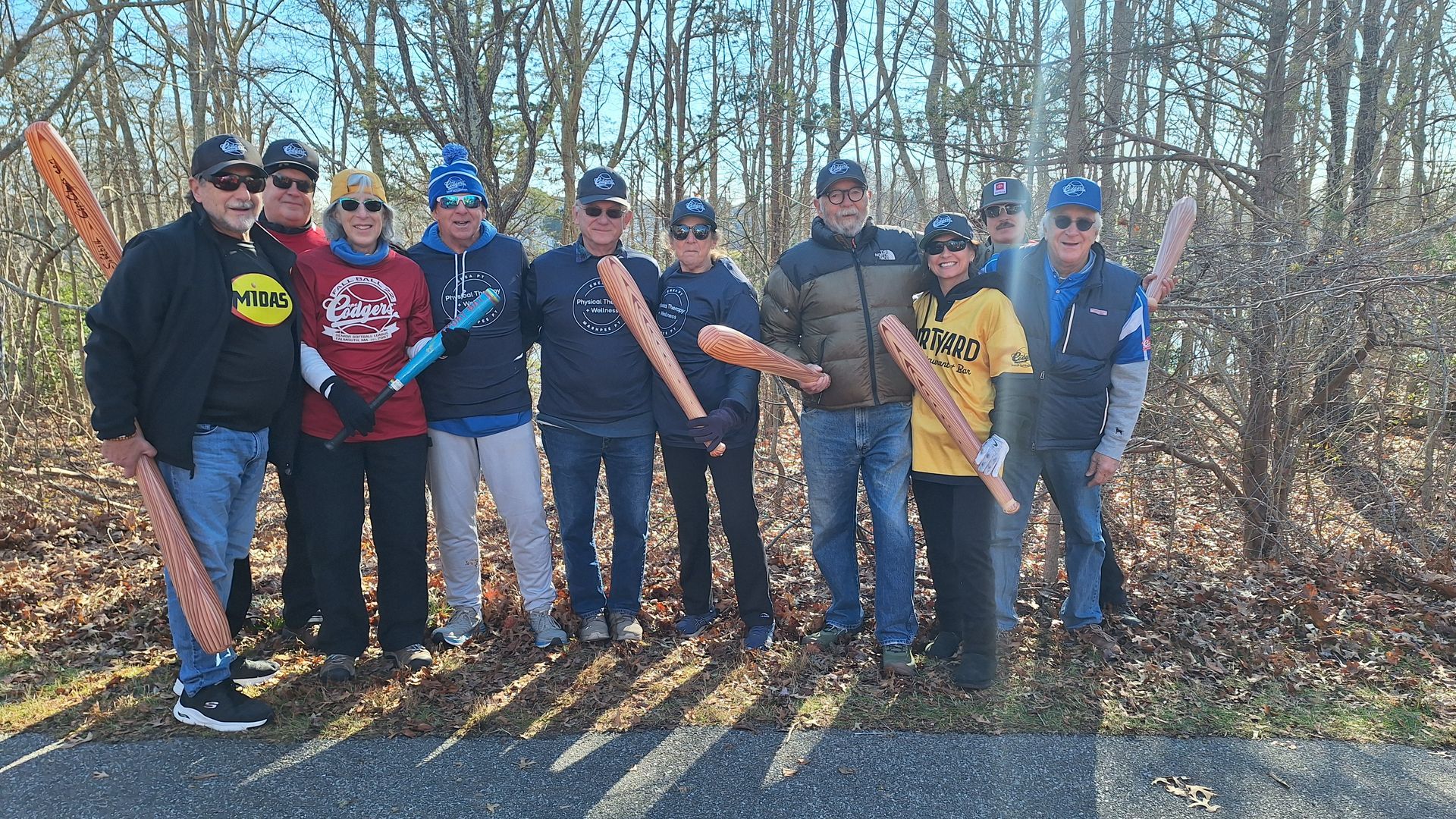 A group of ten people stand in a line outdoors in a wooded area, holding baseball bats and posing for a photo.