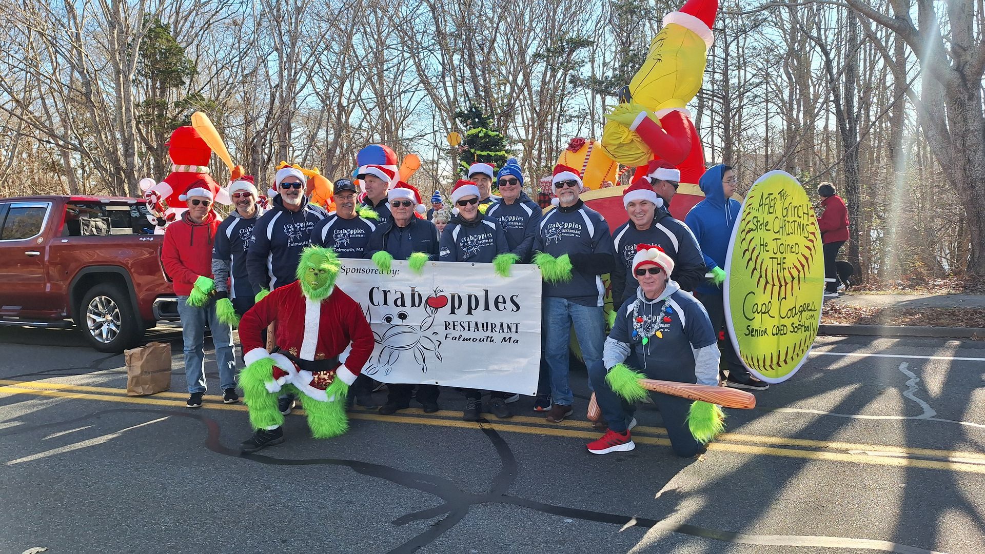 A group in holiday gear, including a Grinch costume, stands behind a Crabapples banner at an outdoor community event.
