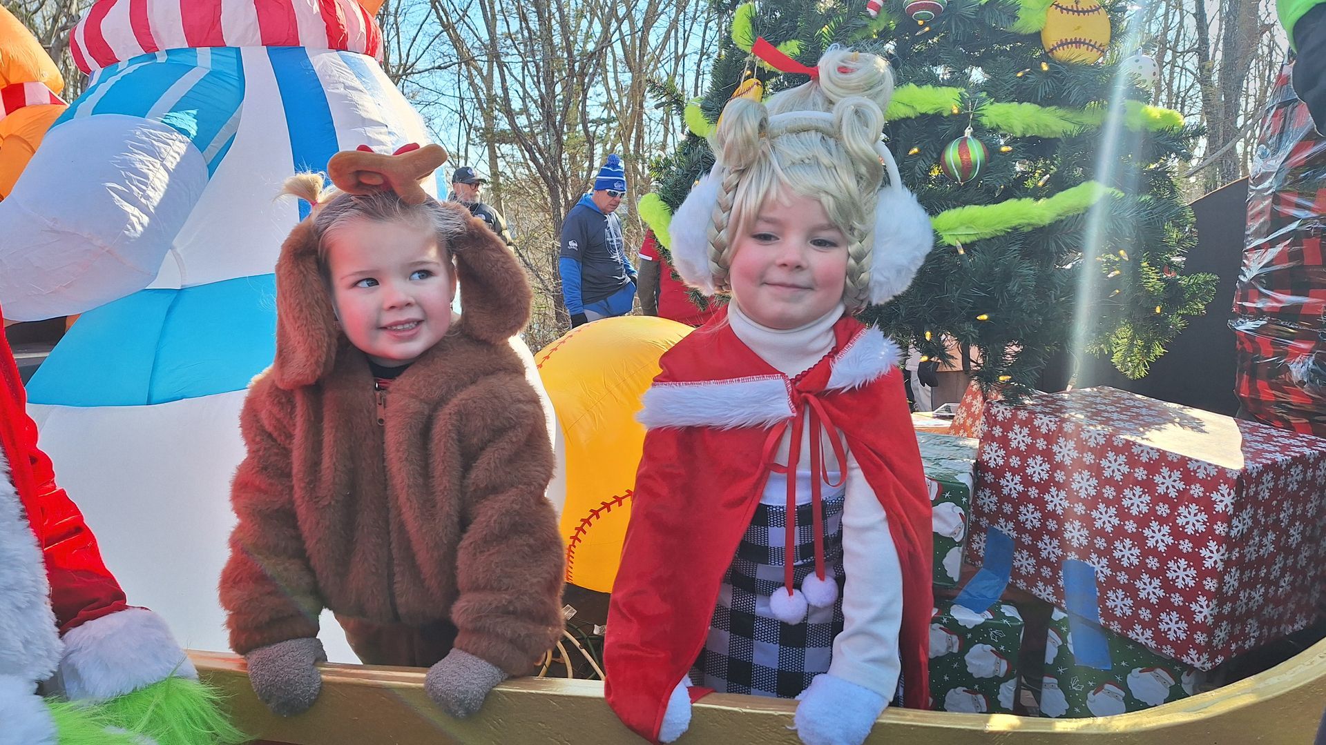 Two children in costumes, one as a reindeer and one as Cindy Lou Who, stand on a parade float decorated for Christmas.