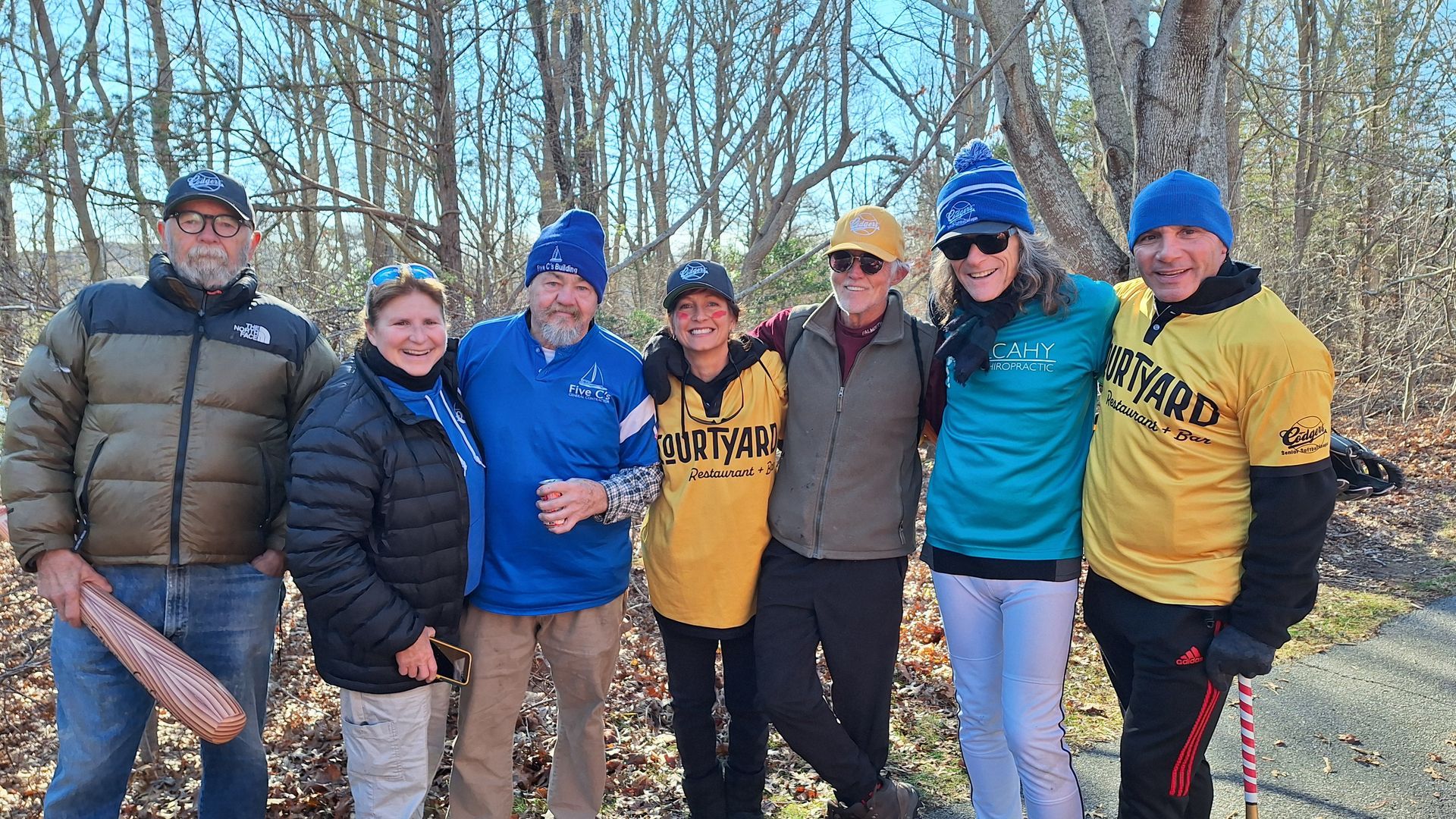 A group of six people stand together outdoors among bare trees, smiling and wearing casual, layered winter clothing.