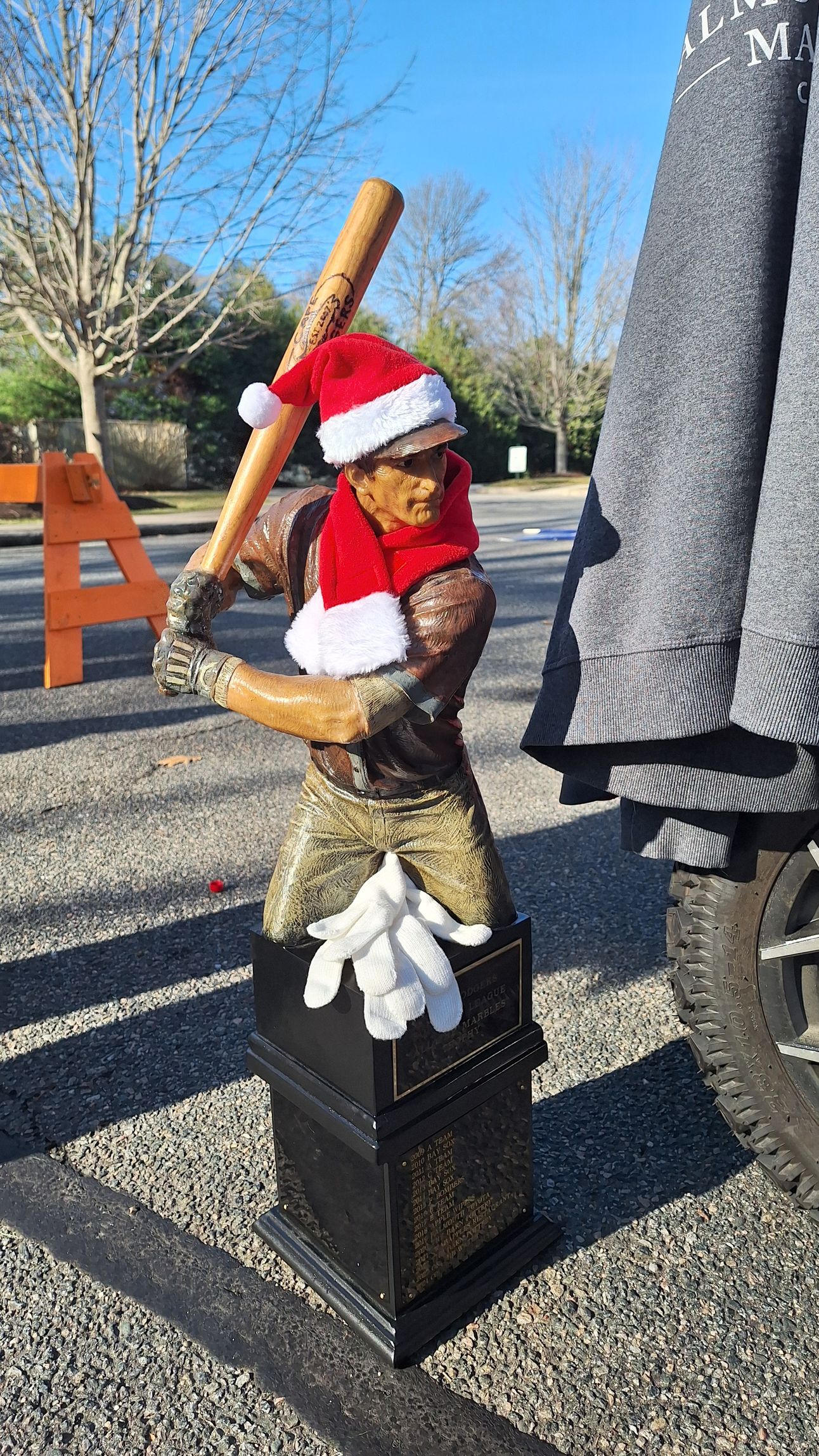 A statue of a baseball player in a Santa hat and red scarf, holding a bat, stands on a black pedestal outdoors.