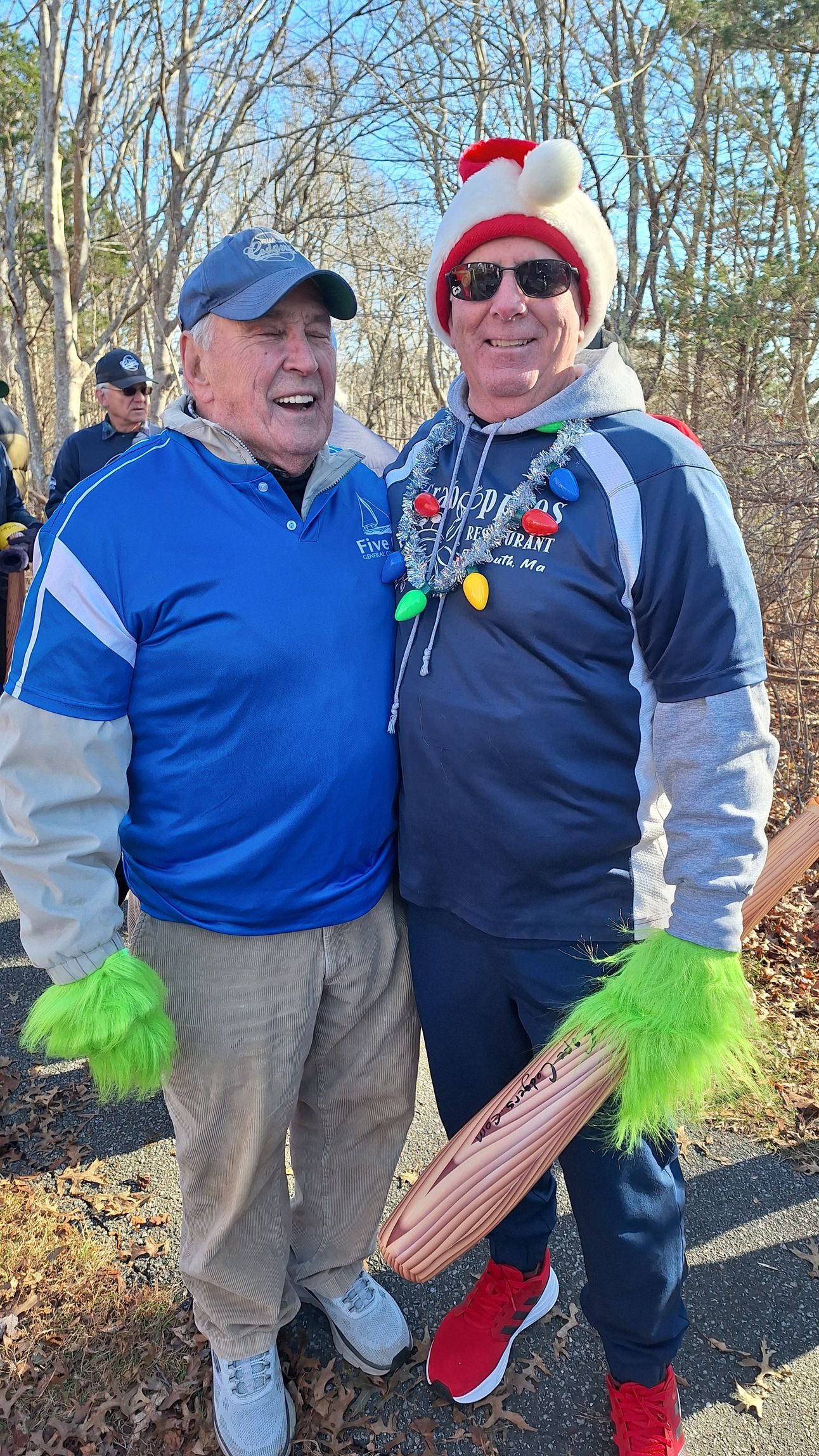 Two people in festive hats and neon green mittens stand smiling outdoors in a wooded area on a sunny day.