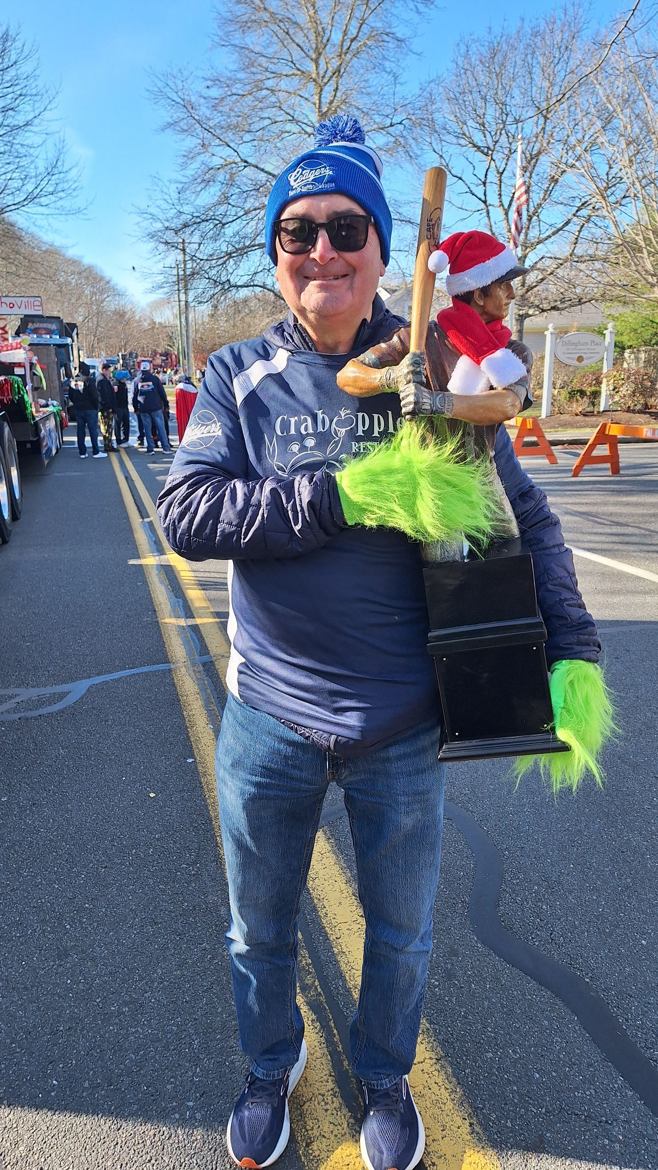 A smiling person wearing a blue beanie and sunglasses stands on a road, holding a small, decorated holiday-themed prop.