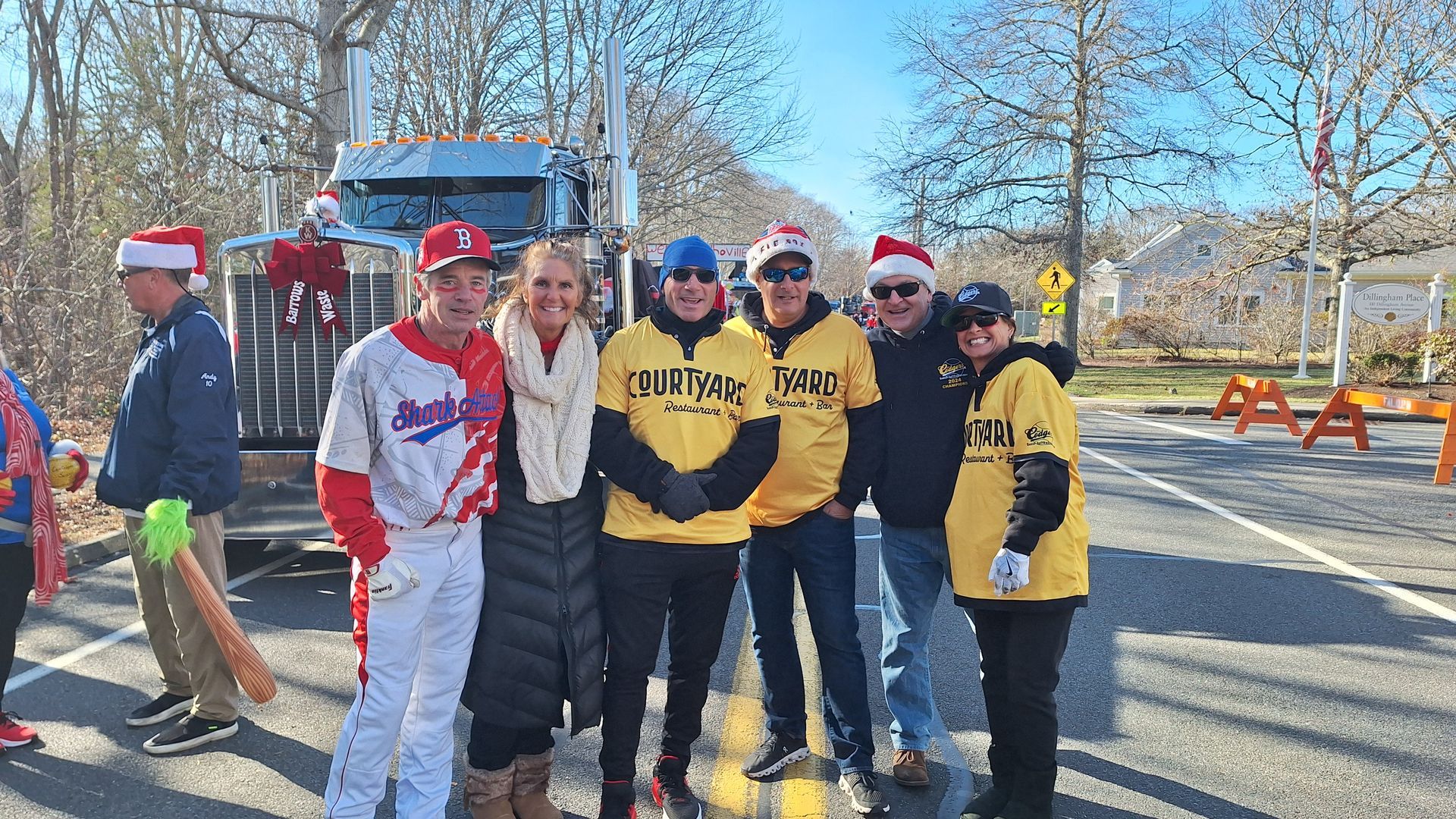 A group of people, some wearing Santa hats and yellow shirts, pose together in front of a parked semi-truck.