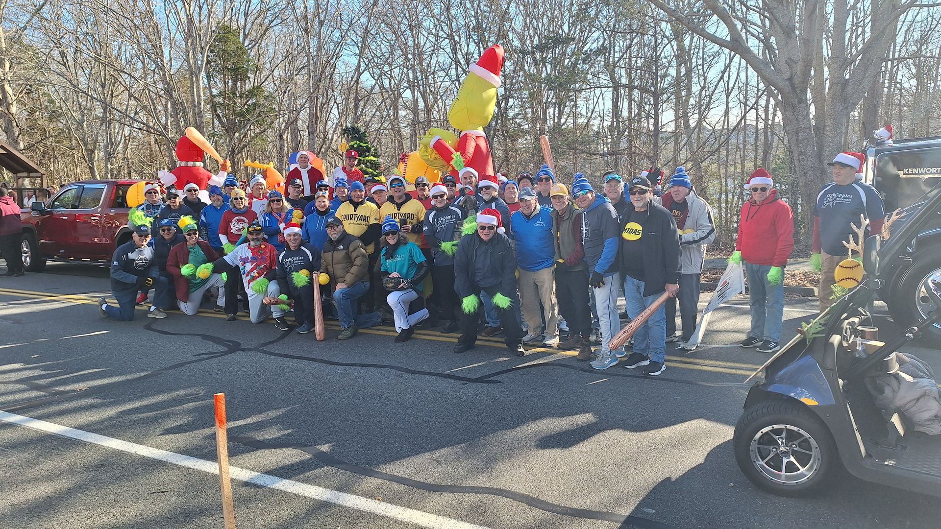 A large group of people in festive attire and Santa hats pose for a photo with holiday inflatables on a sunny parking lot.