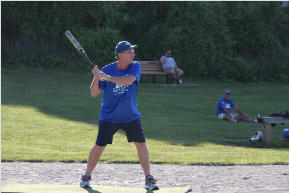 A person in a blue shirt and cap stands on a dirt field, holding a baseball bat in a batting stance.